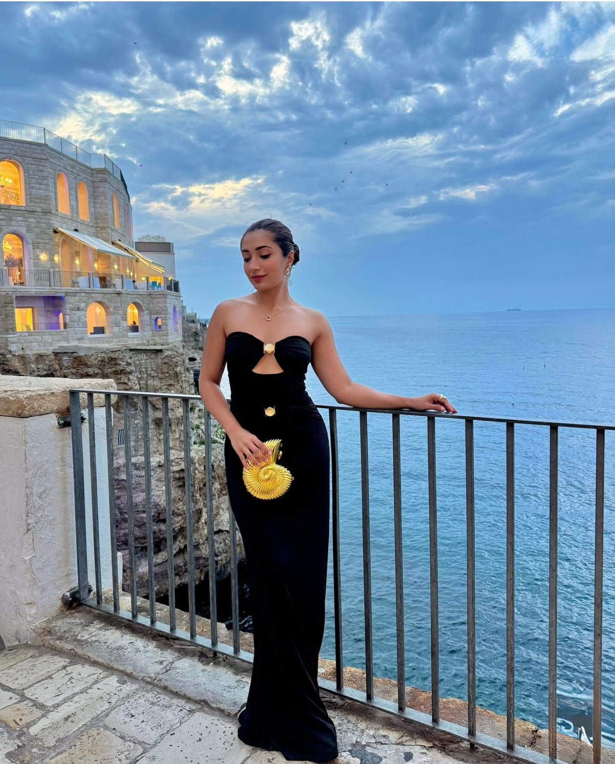 Woman in a black dress with gold accents standing on a balcony overlooking the ocean and a building with golden clutch in Italy